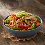 A bowl of Chicken Teriyaki Noodles with sliced chicken, colorful bell peppers, broccoli, carrots, and green onions, topped with sesame seeds.