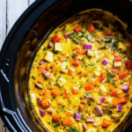 Slow cooker filled with a cheesy breakfast casserole layered with crispy bacon, colorful bell peppers, and golden melted cheddar cheese.