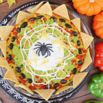Spider Web Taco Dip Recipe with creamy layers of beans, guacamole, cheese, and colorful toppings served with tortilla chips
