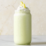 A tall glass of creamy Whipped Lemonade topped with whipped cream and a lemon slice on a wooden table.