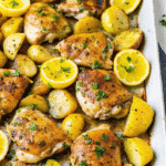 Golden roasted Greek chicken thighs with crispy Yukon potatoes, garnished with fresh parsley and lemon wedges on a sheet pan.