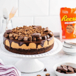 Decadent Reese Cheesecake with brownie base, creamy peanut butter cheesecake layer, chocolate ganache, and mini Reese’s cups on top