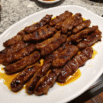 Close-up of golden honey-glazed steak strips sizzling on a hot grill pan, garnished with garlic and spices.