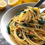 A bowl of spaghetti with garlic and spinach topped with grated Parmesan cheese and lemon wedges on the side.