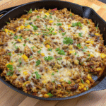 Skillet filled with cheesy taco rice made with ground beef, black beans, corn, tomatoes, and melted Colby Jack cheese topped with sour cream and cilantro.