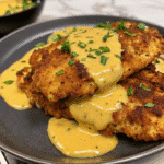 Golden pretzel crusted chicken breast sliced and topped with creamy mustard cheddar sauce on a white plate.