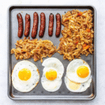 Sheet Pan Breakfast with crispy hash browns, golden baked eggs, breakfast sausage, melted cheese, and fresh herbs on a baking sheet