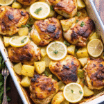 Golden roasted Greek lemon chicken and potatoes with herbs, garlic, and lemon slices in a baking dish.